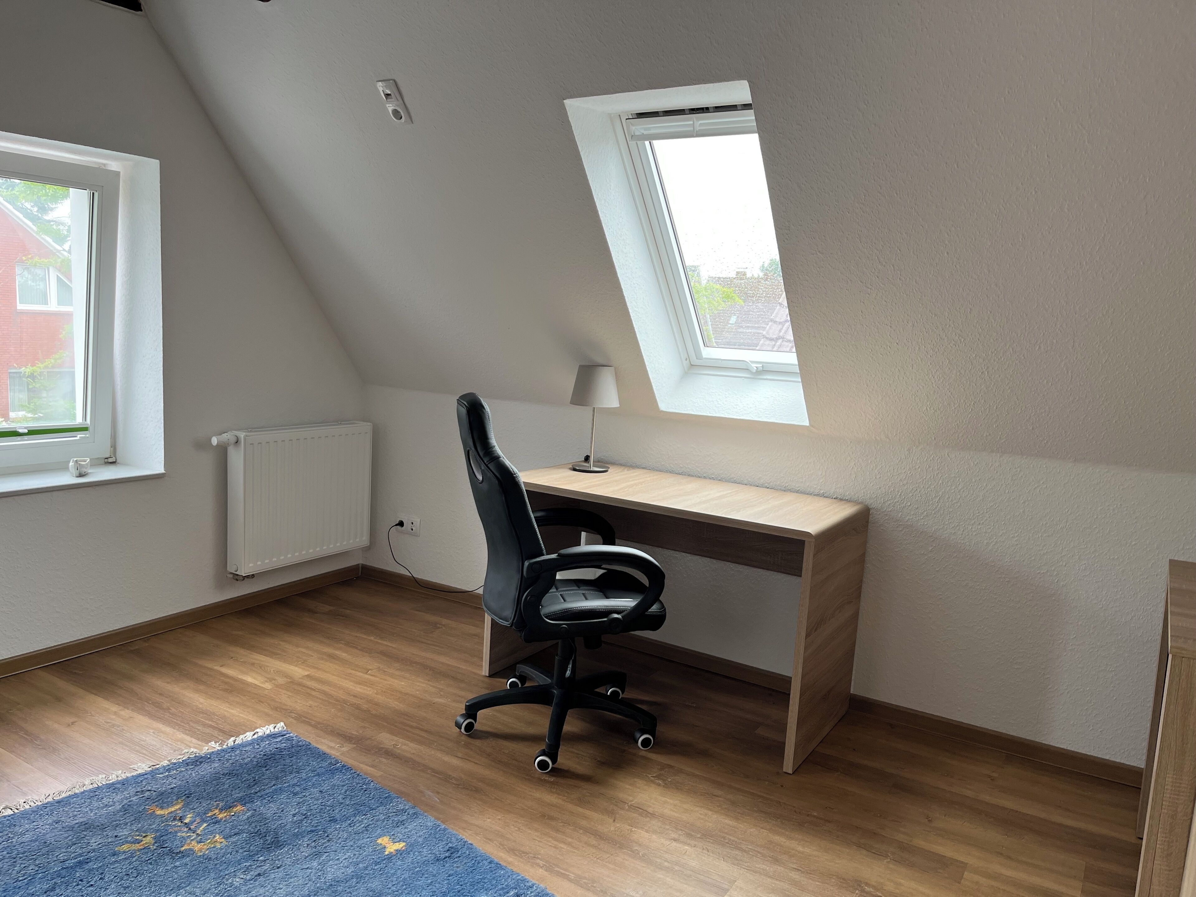 A small, well-lit room with a sloped ceiling, a wooden desk, a black office chair, a small table lamp, a blue rug, and a window with a view of a house.