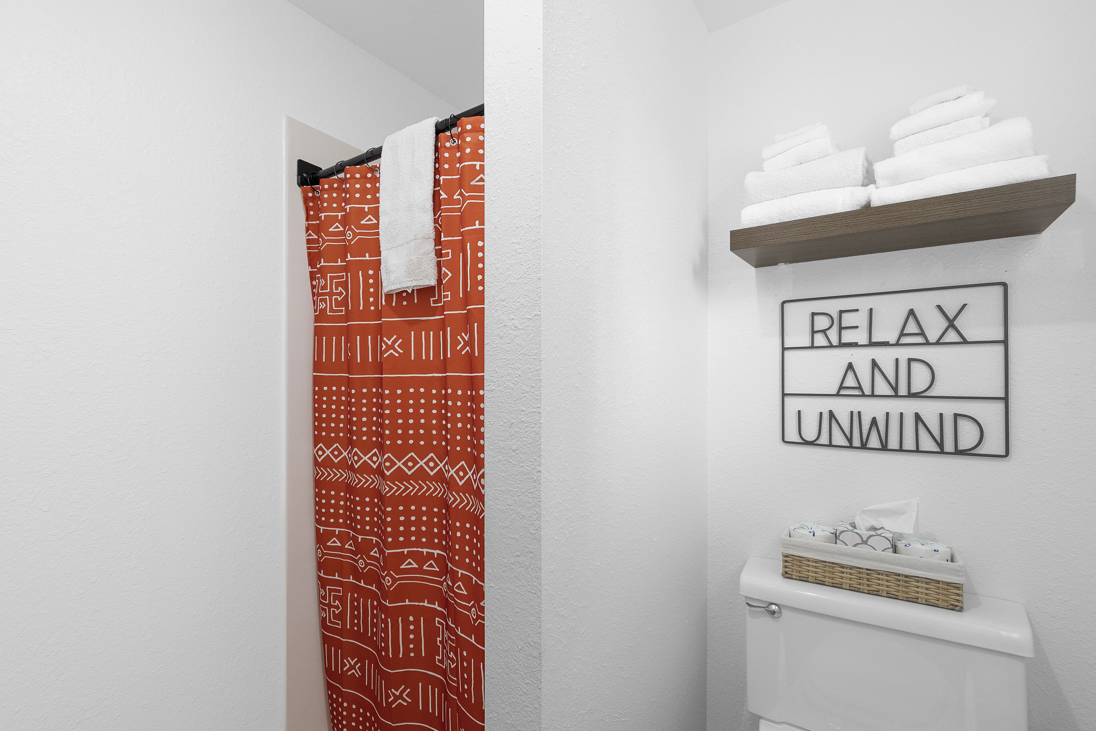 A bathroom with a patterned shower curtain, a towel rack with folded towels, and a sign that reads "RELAX AND UNWIND".