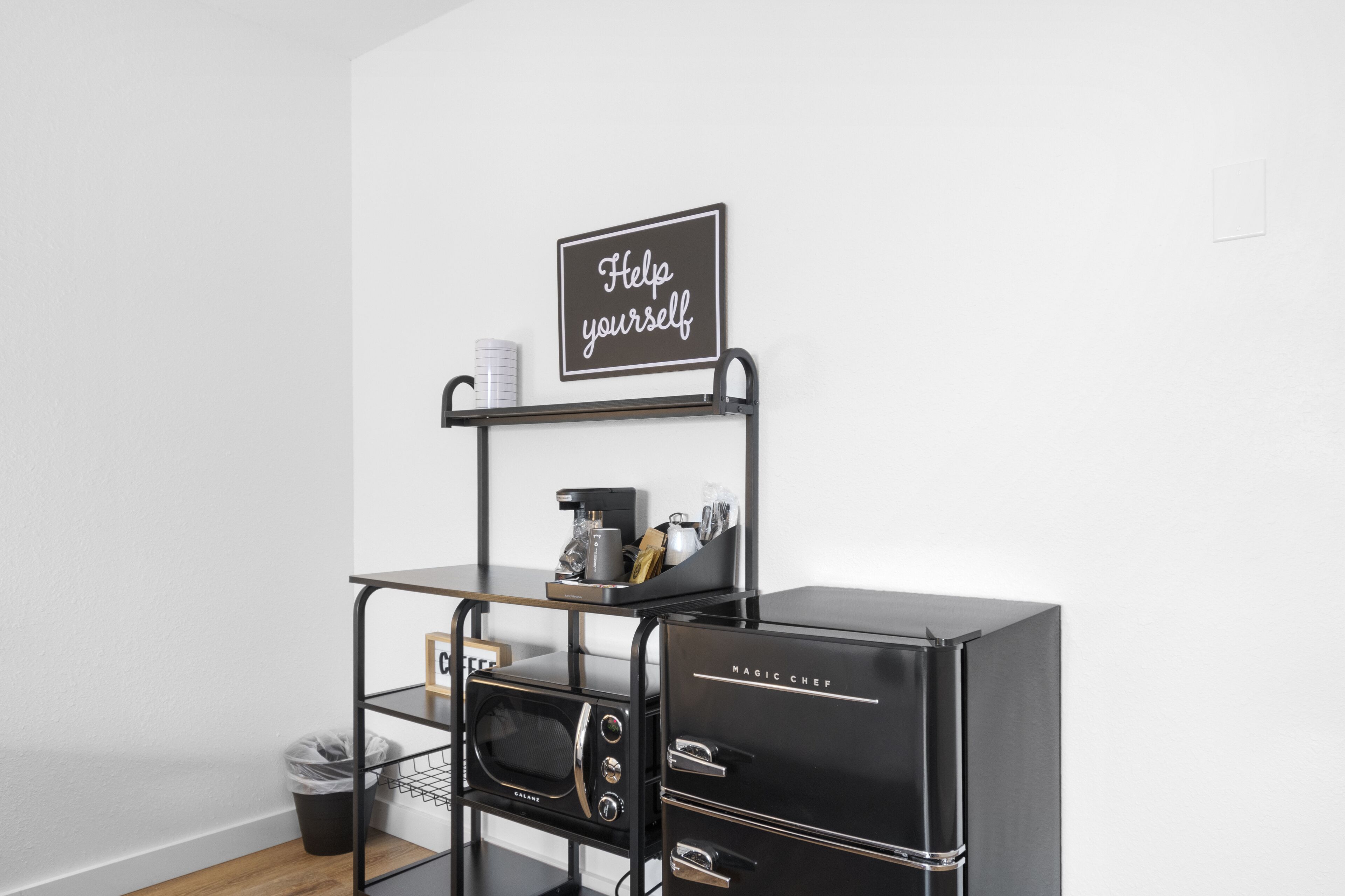 Un área de cocina con un refrigerador negro, una cafetera y un letrero que dice 'Sírvaste a tu gusto'.