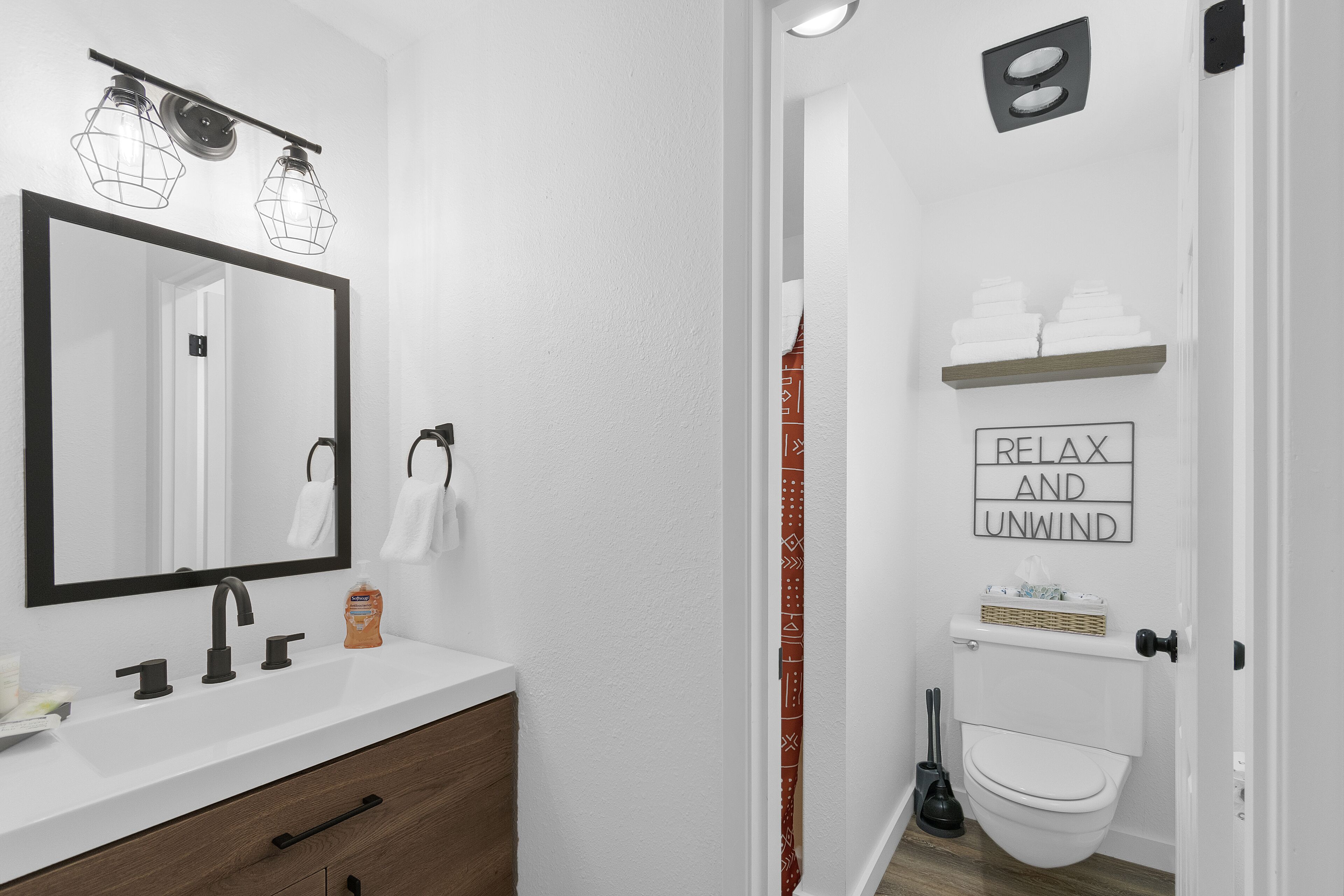 A modern bathroom with a large mirror, a white sink, wooden cabinet, and a toilet. There is a sign that says "RELAX AND UNWIND" on the wall.