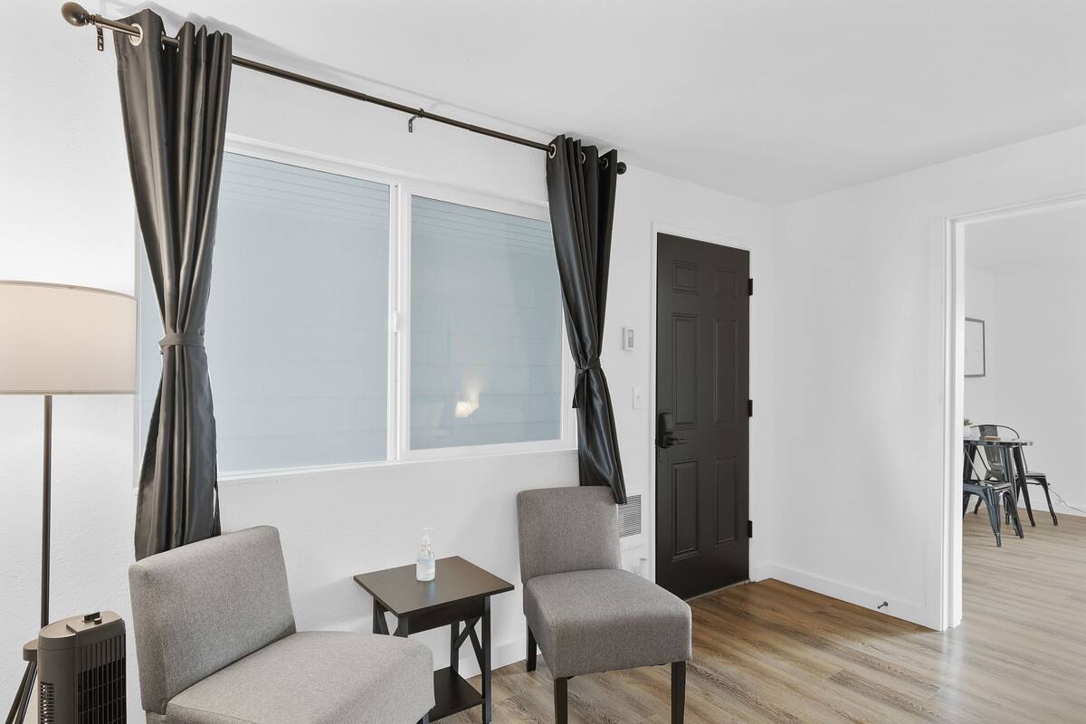 Quarto com duas poltronas cinzentas, uma mesa pequena, um abajur e uma janela com cortinas.