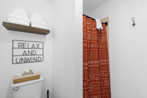 A bathroom with a toilet, a shelf with towels, a sign that reads "RELAX AND UNWIND", and a shower curtain with a geometric pattern. - Silver Sands Motel (Westport)