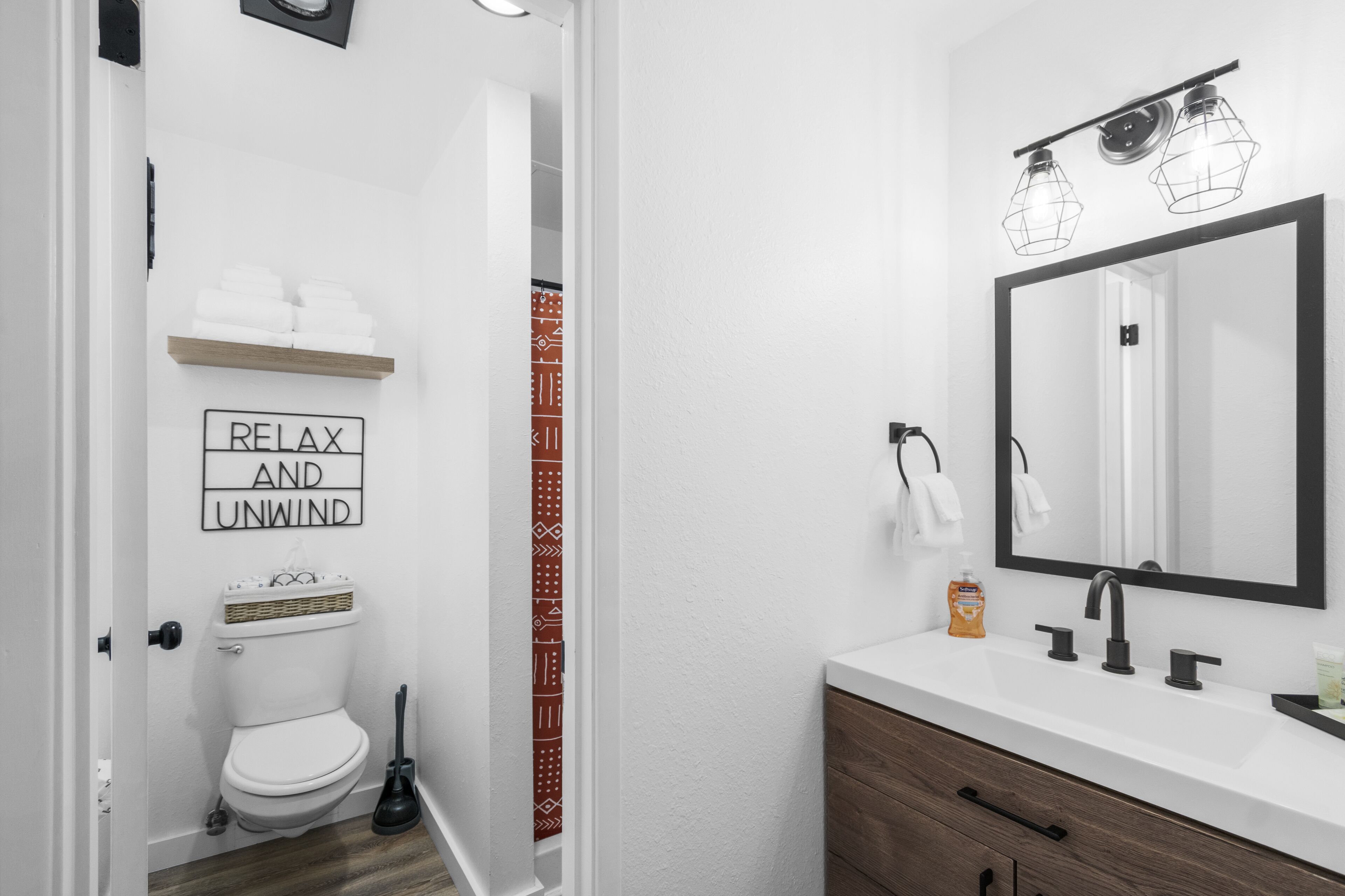 Baño con inodoro, lavamanos y espejo. Hay una repisa con toallas y un cartel que dice "RELAX AND UNWIND".