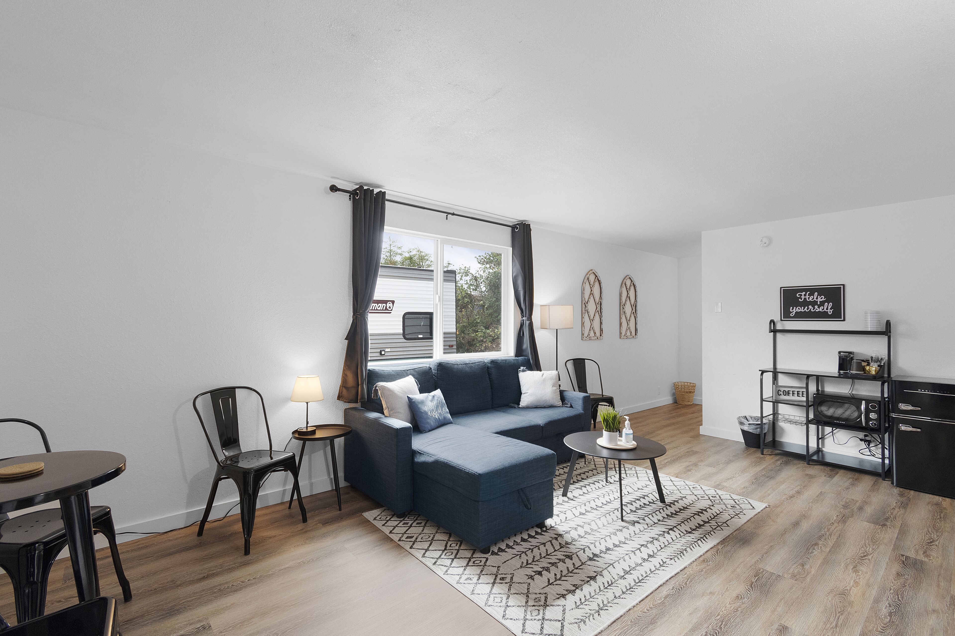 A modern living room with a blue sectional sofa, a dining table, chairs, a coffee station, and a view of the outside.