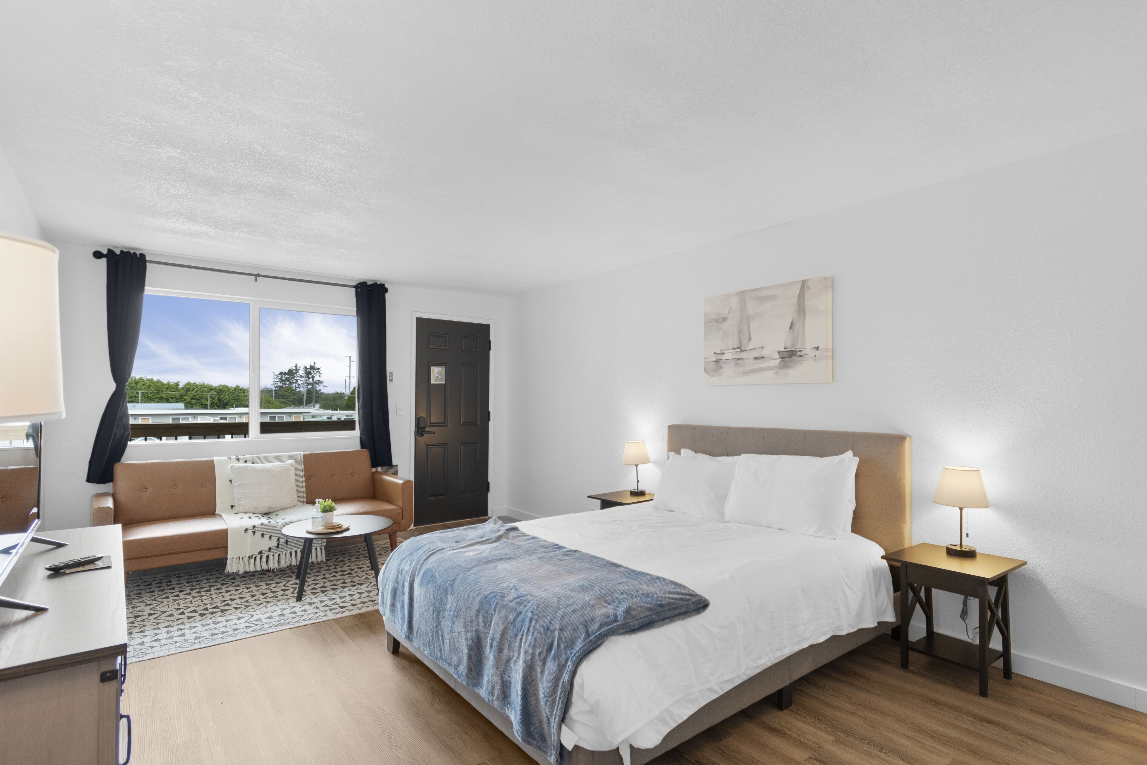 A hotel room with a bed, a sofa, a small table, and a desk. There is a window with curtains, a door, and a painting on the wall.
