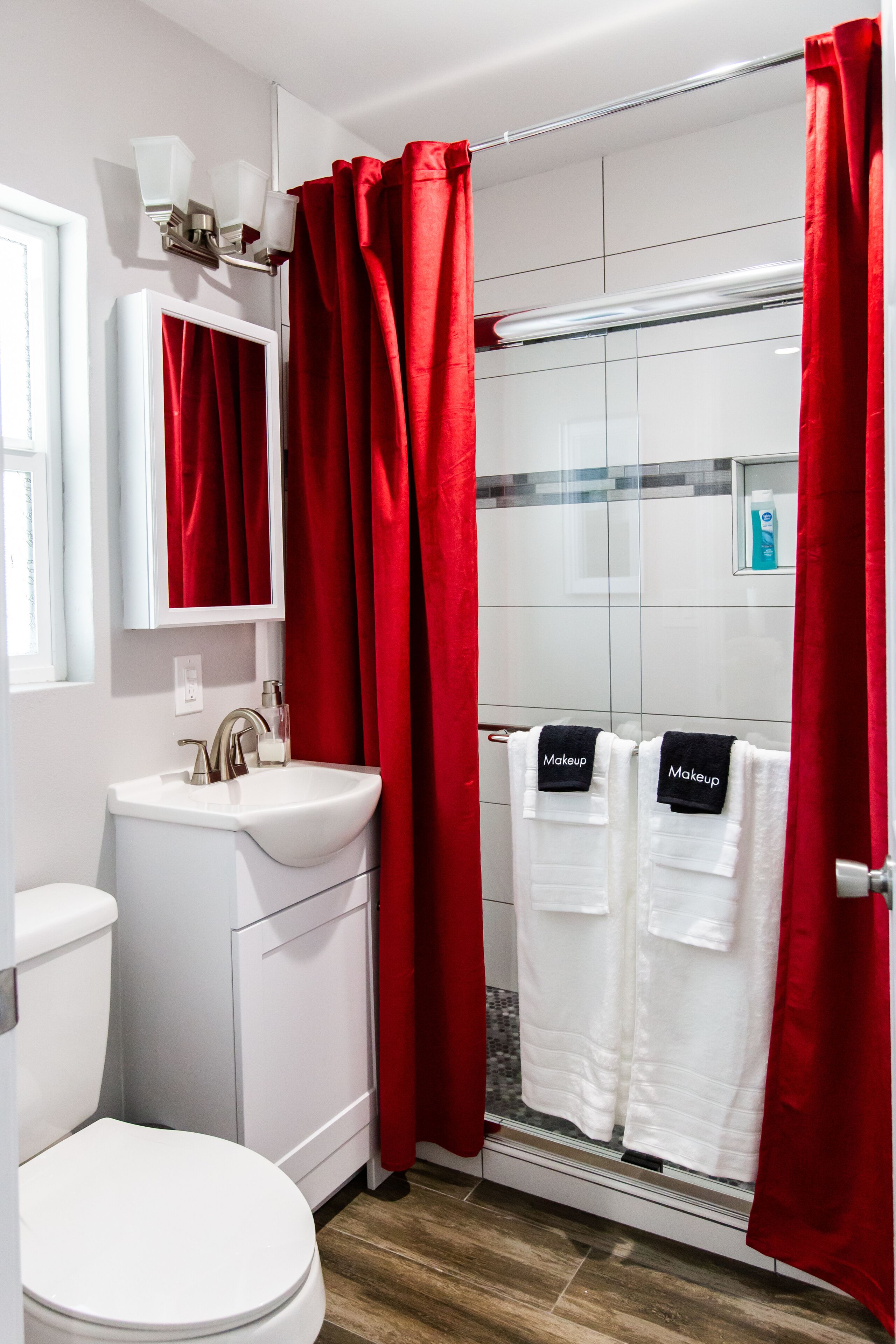 Un bagno con lavandino bianco, un water e zona doccia con tendaggi rossi.