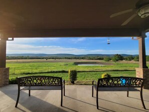 A covered outdoor seating area with two ornate metal benches facing a scenic view of a green field and distant hills.