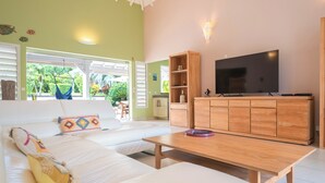 Un salon comprenant un canapé, une table basse en bois, un téléviseur et un meuble en bois. Une fenêtre offre une vue sur la verdure à l’extérieur.