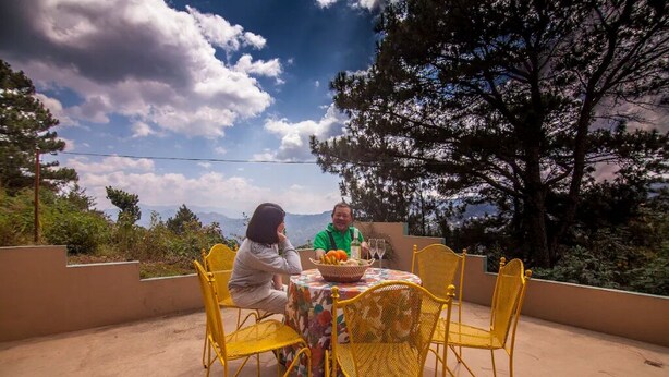 Zwei Personen sitzen an einem Tisch mit einer Obstschale, umgeben von gelben Stühlen, mit Blick auf Bäume und Berge im Hintergrund.