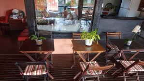 A cozy outdoor seating area with wooden tables and chairs, potted plants, and a glass partition. - Pousada Casa da Tatá (Búzios)