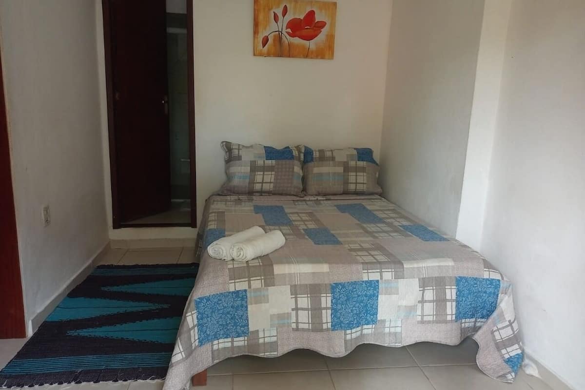 A bedroom with a bed, a blue and beige checkered bedspread, a blue and beige pillow, a red flower painting on the wall, and a blue and black area rug.