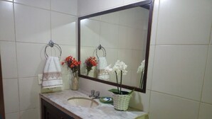 A bathroom with a sink, mirror, and two towels hanging on towel rings. There are two flower arrangements on the sink counter. - Pousada Casa da Tatá (Búzios)
