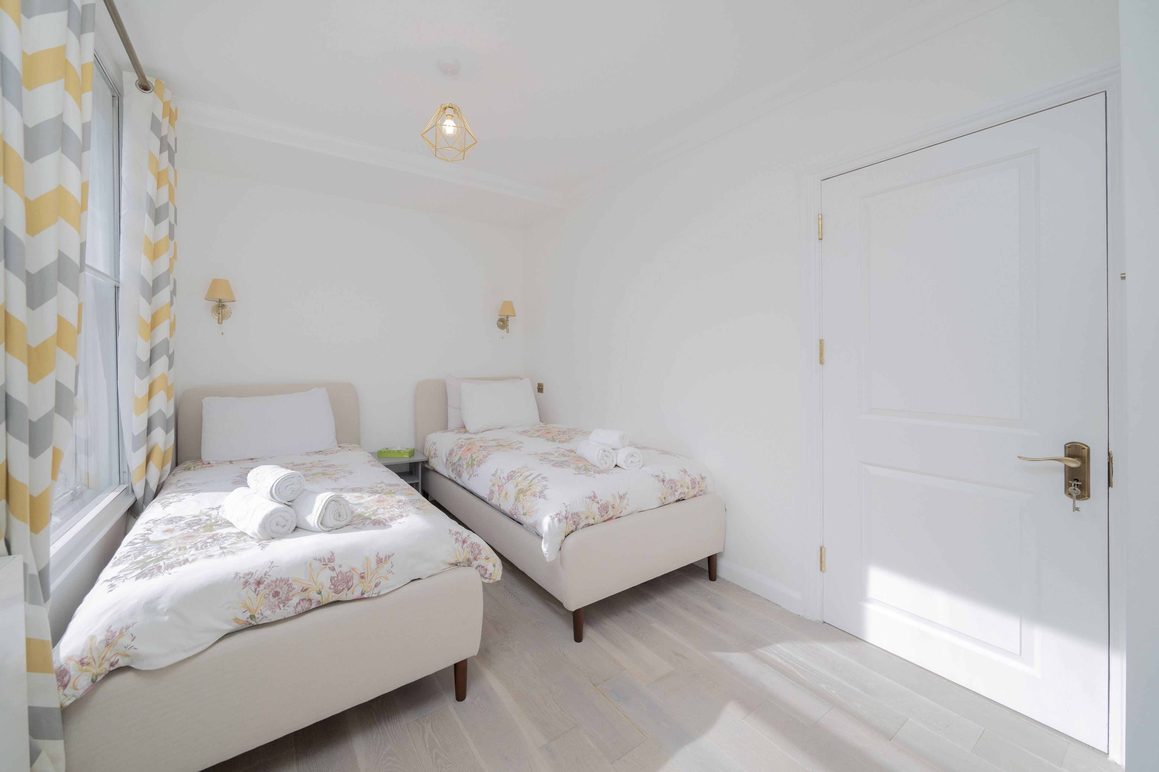 A bedroom with two beds, a window with curtains, a door, and a light fixture.