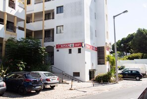 A multi-story building with a white facade, a visible entrance, and a staircase leading to the ground floor. There are parked cars in front, and a street lamp is visible to the right.