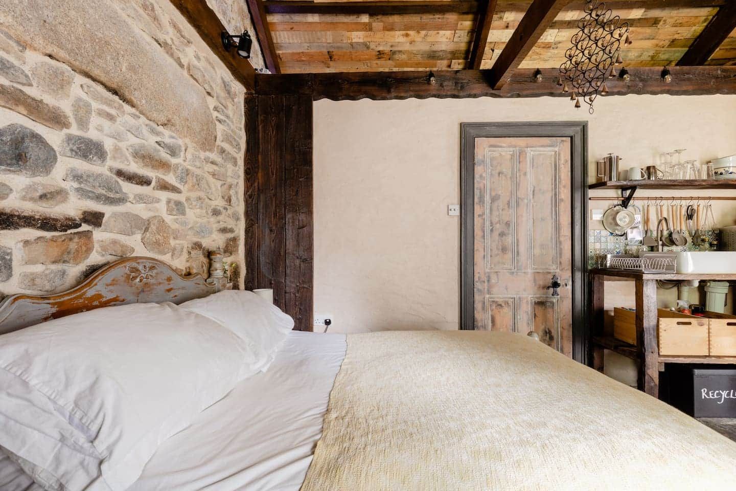 Un dormitorio rústico con pared de piedra, vigas de madera, una cama con ropa de cama blanca, una puerta de madera y una zona de cocina con utensilios colgando.