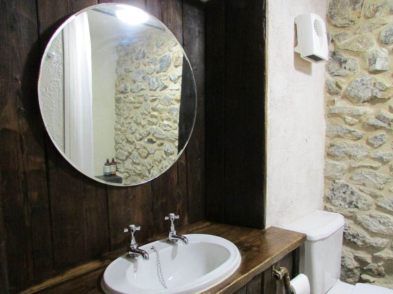 Un baño con un espejo redondo, dos lavamanos, un inodoro y una pared de piedra.
