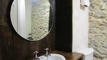 Un baño con un espejo redondo, dos lavamanos, un inodoro y una pared de piedra.