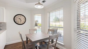 A dining area with a wooden table and chairs, a wall clock, and large windows with blinds. - Home Pool 15 Minutes from DFW Airport (Flower Mound)