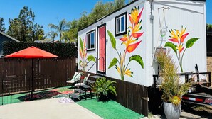 Une petite maison mobile blanche, ornée de peintures murales aux couleurs vives représentant des fleurs sur les côtés, avec une porte rouge et un petit porche aménagé avec du mobilier d’extérieur.