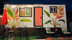 Une petite maison avec une porte rouge, une fenêtre et un carillon. Il y a une chaise et une table à l’extérieur.