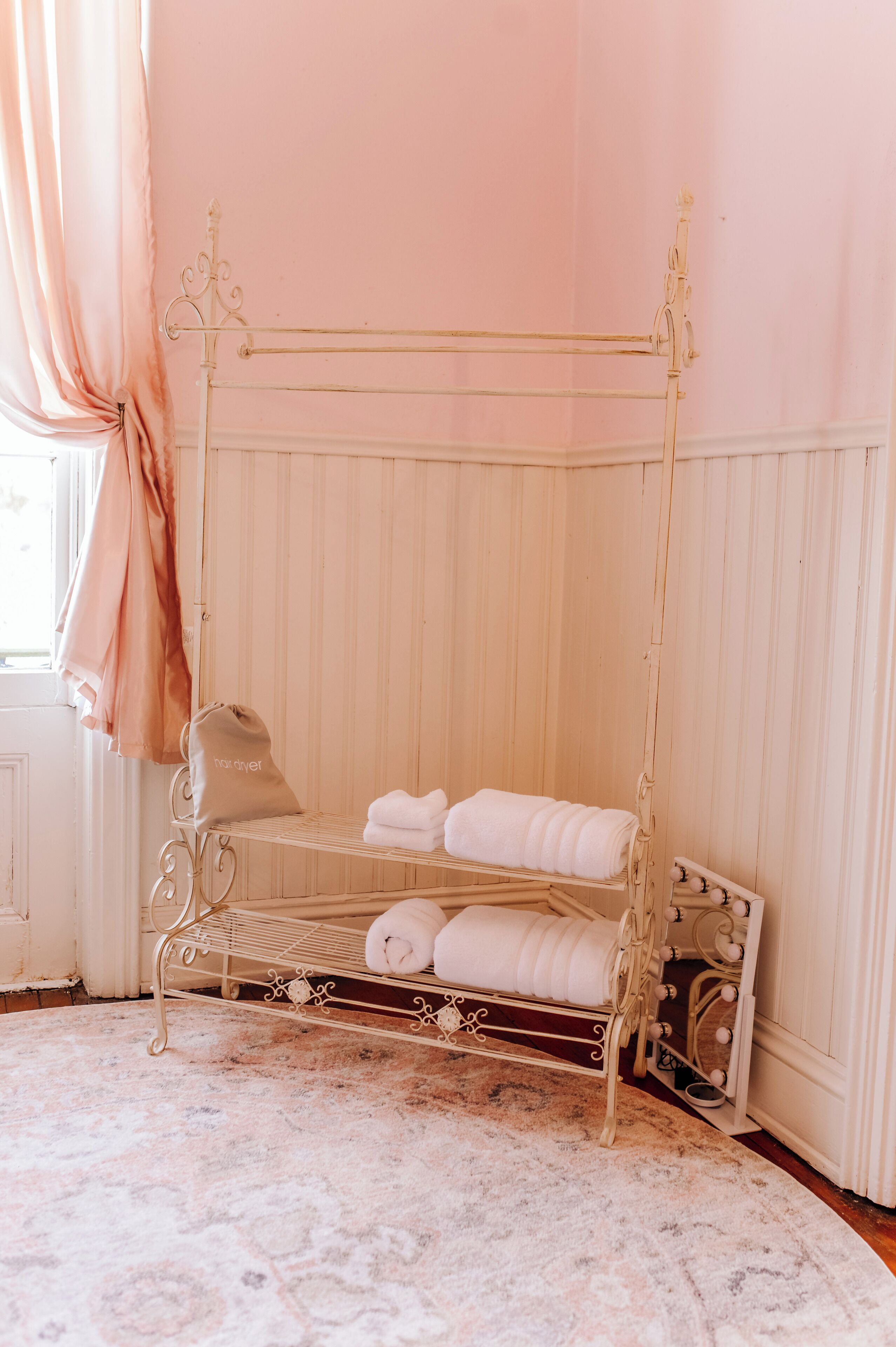 A four-poster bed with white linens and a beige bag on a patterned rug in a room with pink walls and a window with curtains.