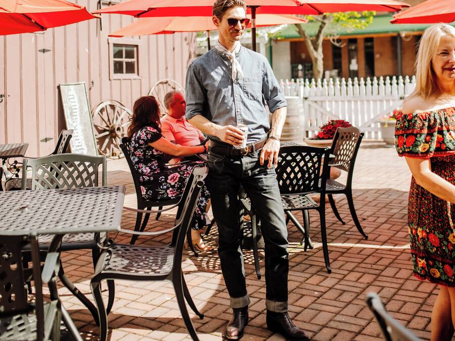 An outdoor seating area with tables and chairs, red umbrellas, and a brick building in the background.