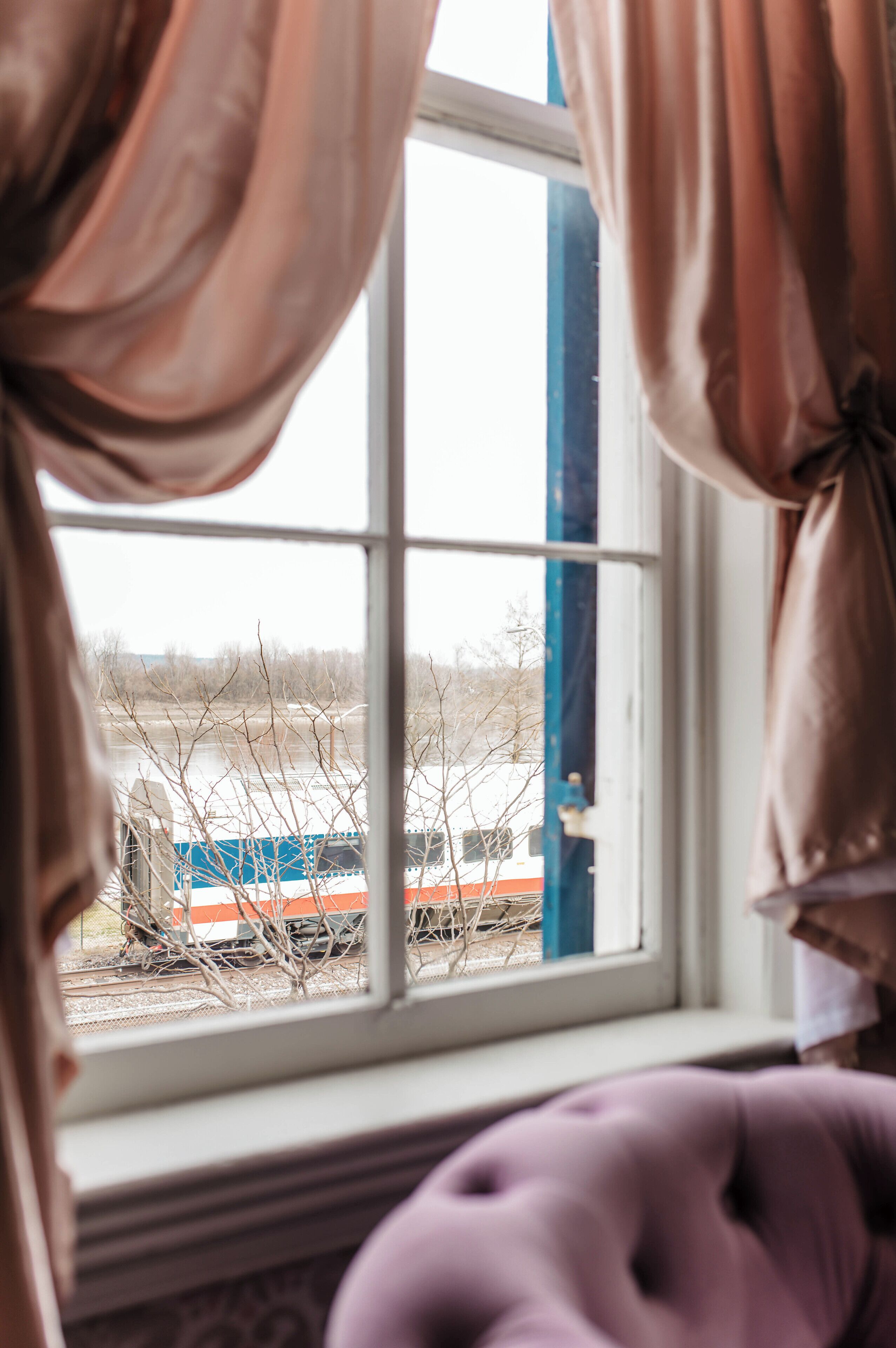 A window with curtains, a view of a train, and a purple chair.
