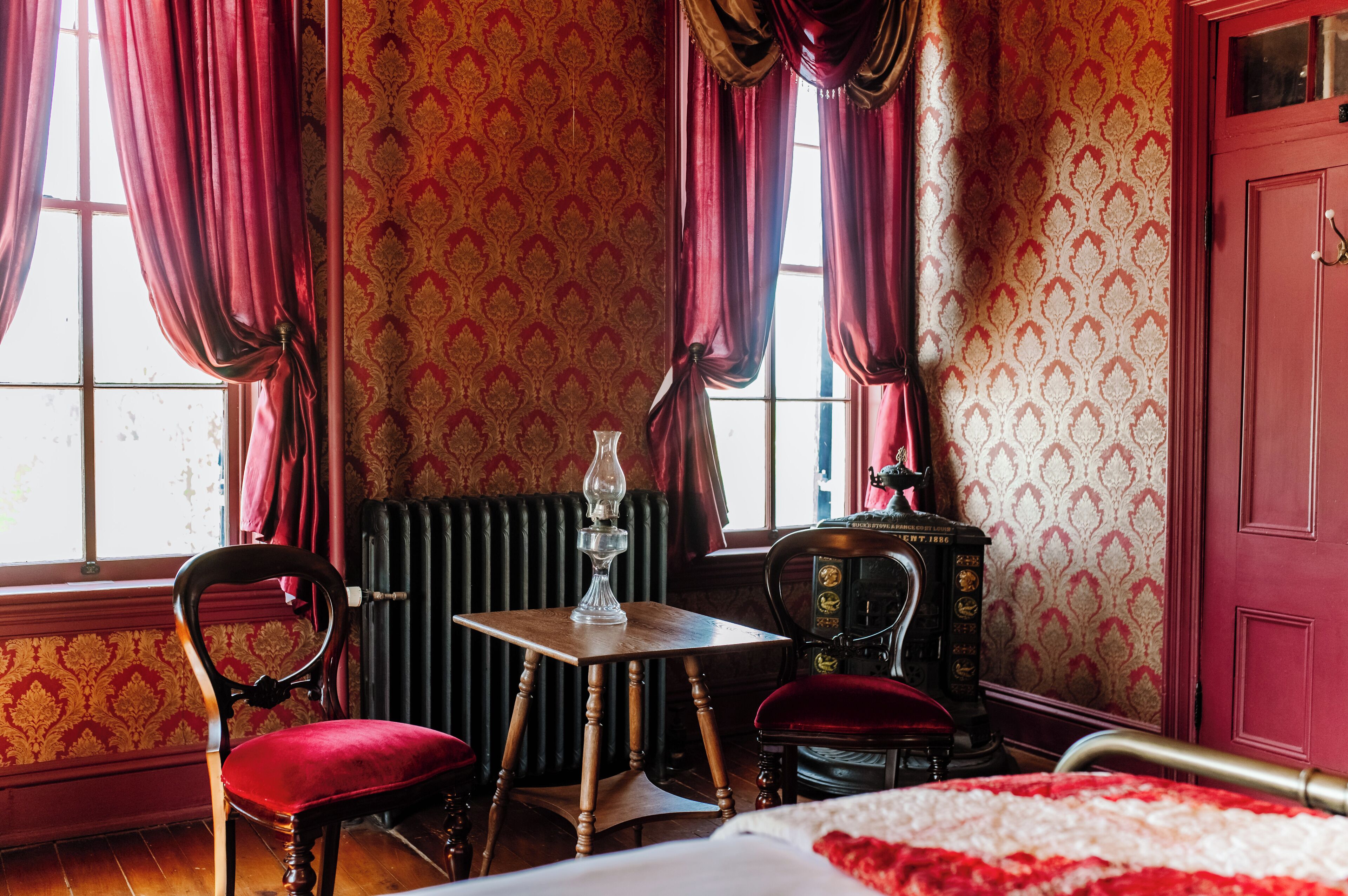 Een kamer met een houten tafel, twee stoelen, een radiator en een bed met een rood-witte dekbedovertrek.