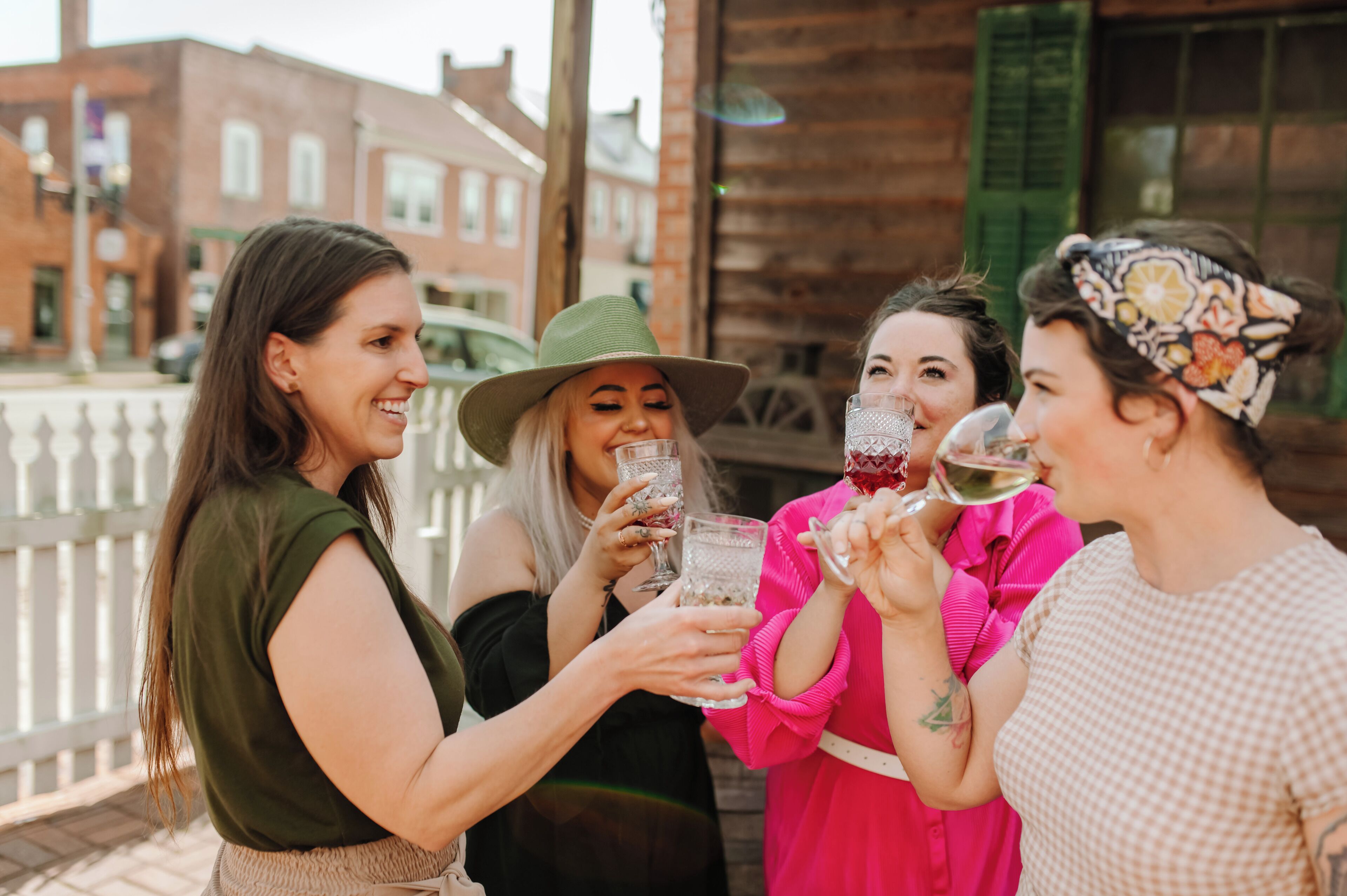 A group of people toasting with drinks on a wooden deck with a white fence and brick buildings in the background.