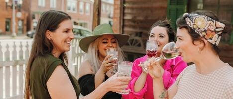 A group of people toasting with drinks on a wooden deck with a white fence and brick buildings in the background.