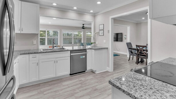 A modern kitchen with white cabinets, granite countertops, and a dishwasher. There is a dining area with a table and chairs, and a ceiling fan.
