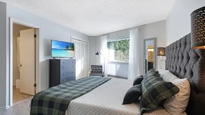 A bedroom with a bed, a television, a dresser, and a window with a view of greenery.