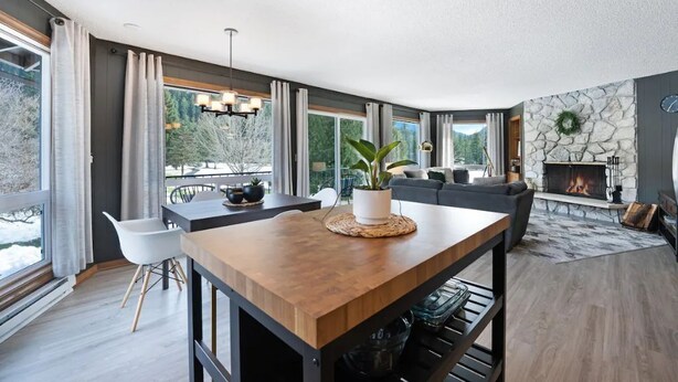 A modern living room with a fireplace, a dining area with a wooden table, and large windows offering a view of the outdoors.