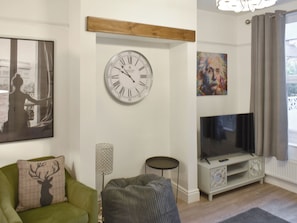 A living room with a large wall clock, a flat-screen TV, a green armchair, a grey ottoman, and a painting. - 4 bedroom accommodation in Keswick (Keswick)