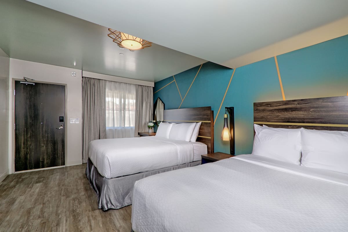 A hotel room with two beds, a wooden door, a curtain, a wall with geometric patterns, and a ceiling light fixture.