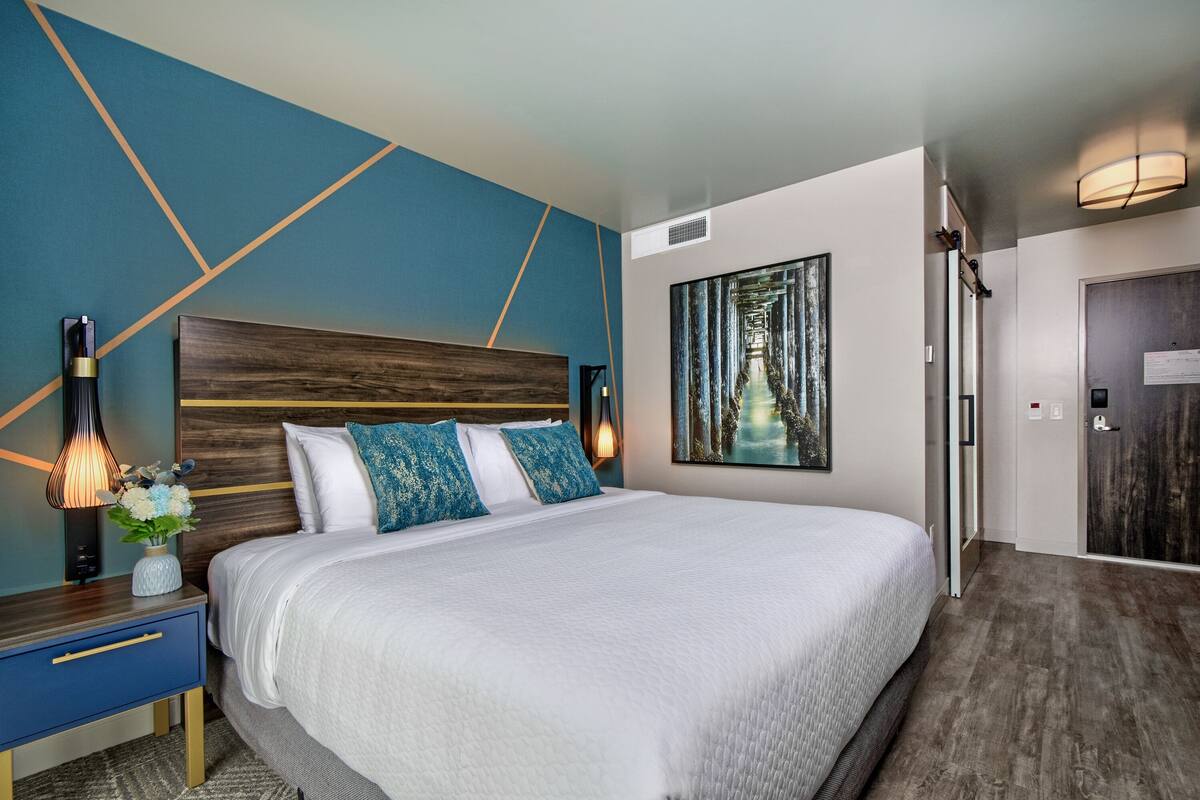 A modern hotel room with a large bed, a nightstand with a lamp, and a painting on the wall.