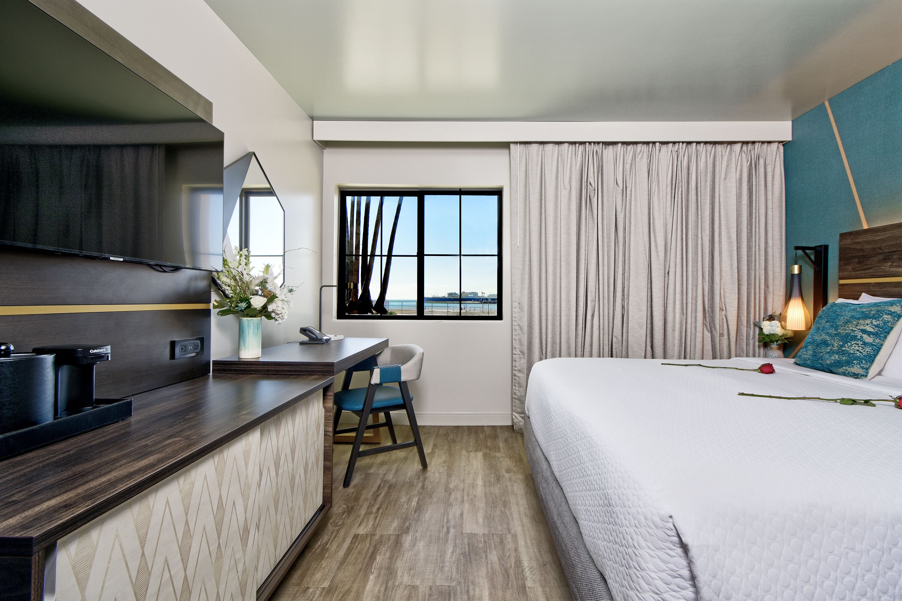 Habitación de hotel con cama, un escritorio con silla, televisor, ventana con cortinas y vista a la ciudad.