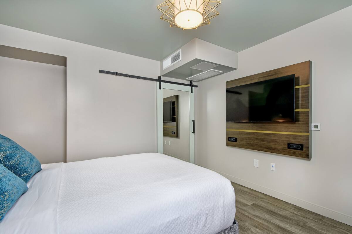 A modern hotel room with a bed, a mounted TV, a ceiling light fixture, and a door with a mirror.