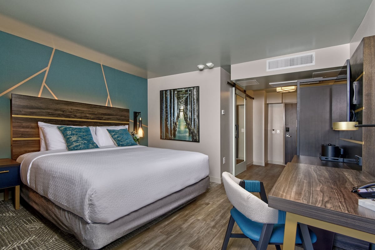 A modern hotel room with a large bed, a desk, and a view into the kitchen.