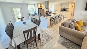 Una cocina moderna con una isla central, comedor con mesa y sillas, y sala de estar con sofá y cojines.