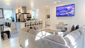 Una sala de estar moderna con un sofá gris, un comedor con mesa y sillas, una cocina con armarios y electrodomésticos blancos, y un televisor de pantalla plana montado en la pared.