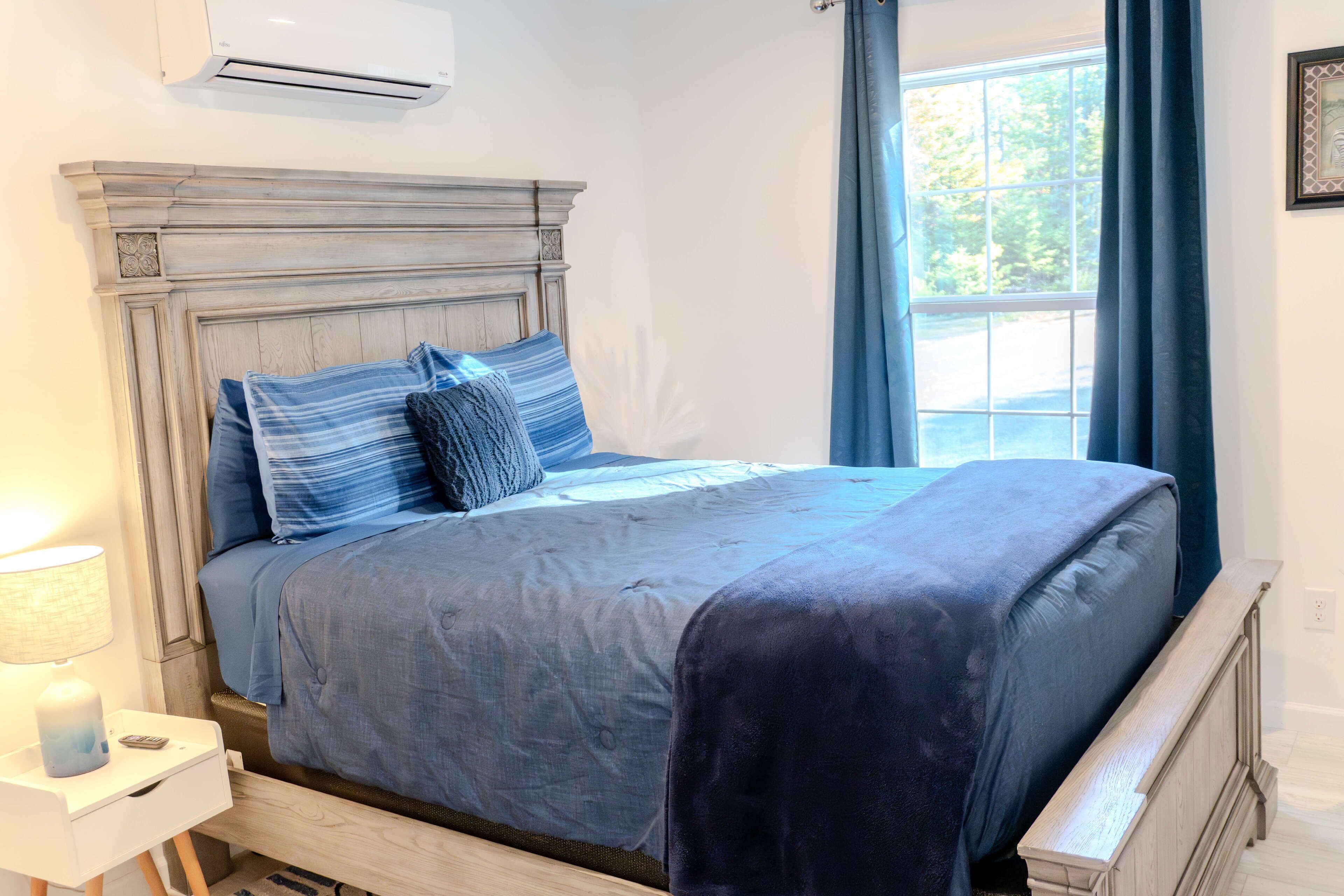 A bedroom with a wooden headboard, a bed with blue bedding, a nightstand with a lamp, and a window with curtains.