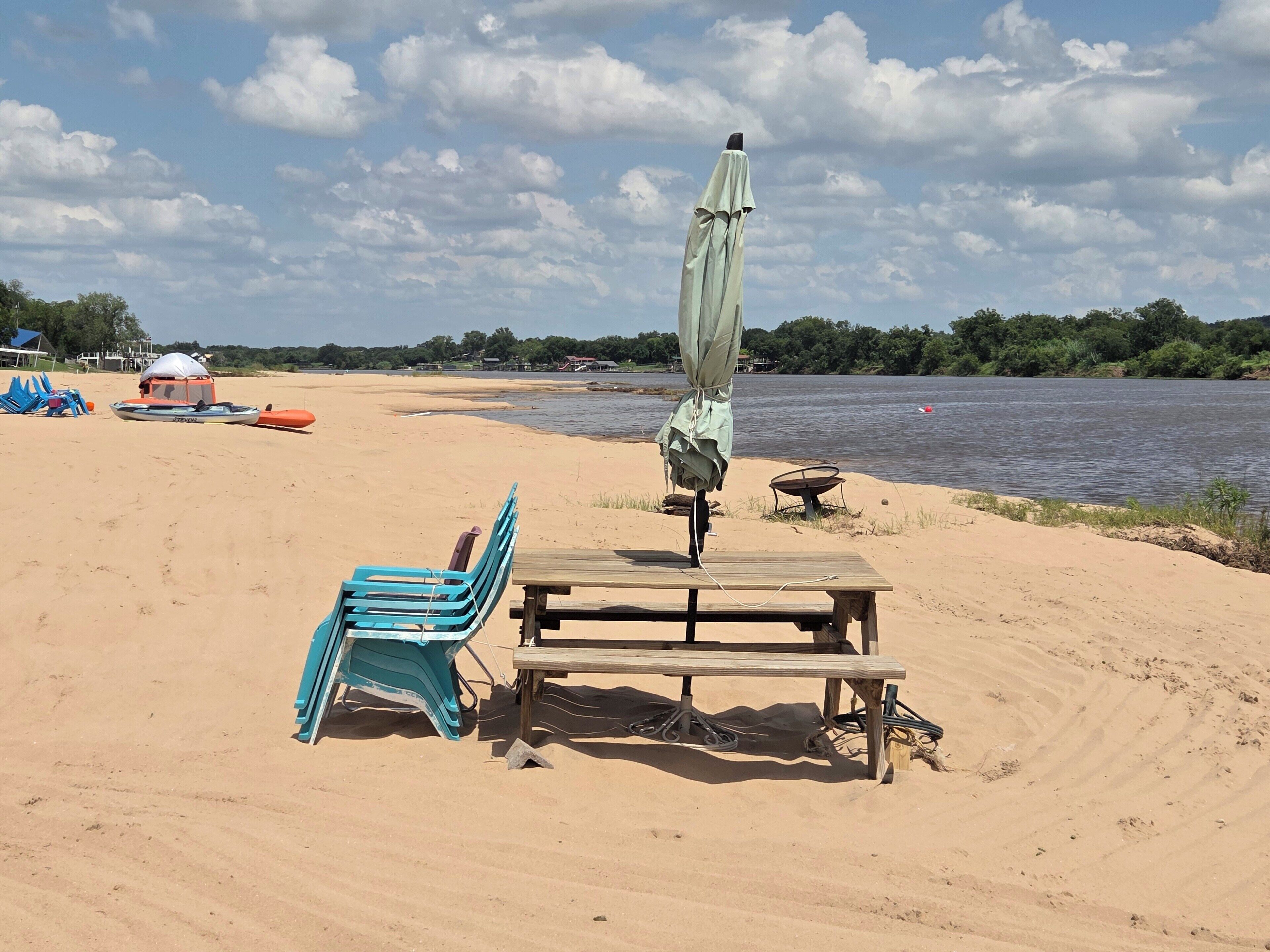 Una playa arenosa con una mesa y sillas, una sombrilla y un barco al fondo.