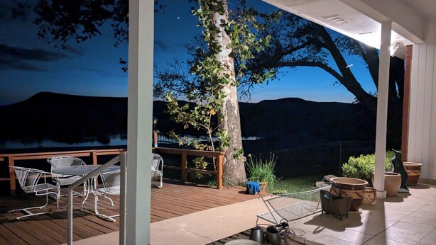 Una terraza con mobiliario exterior, una mesa con un tarro y un vaso, y vistas a un árbol y colinas.