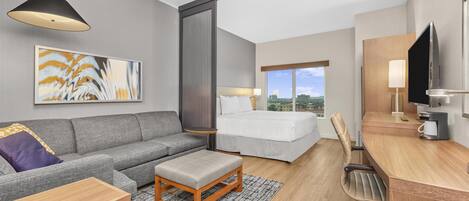 Ett modernt hotellrum med en grå soffa, en vit säng, ett skrivbord i trä, en tv och ett stort fönster med utsikt över staden.