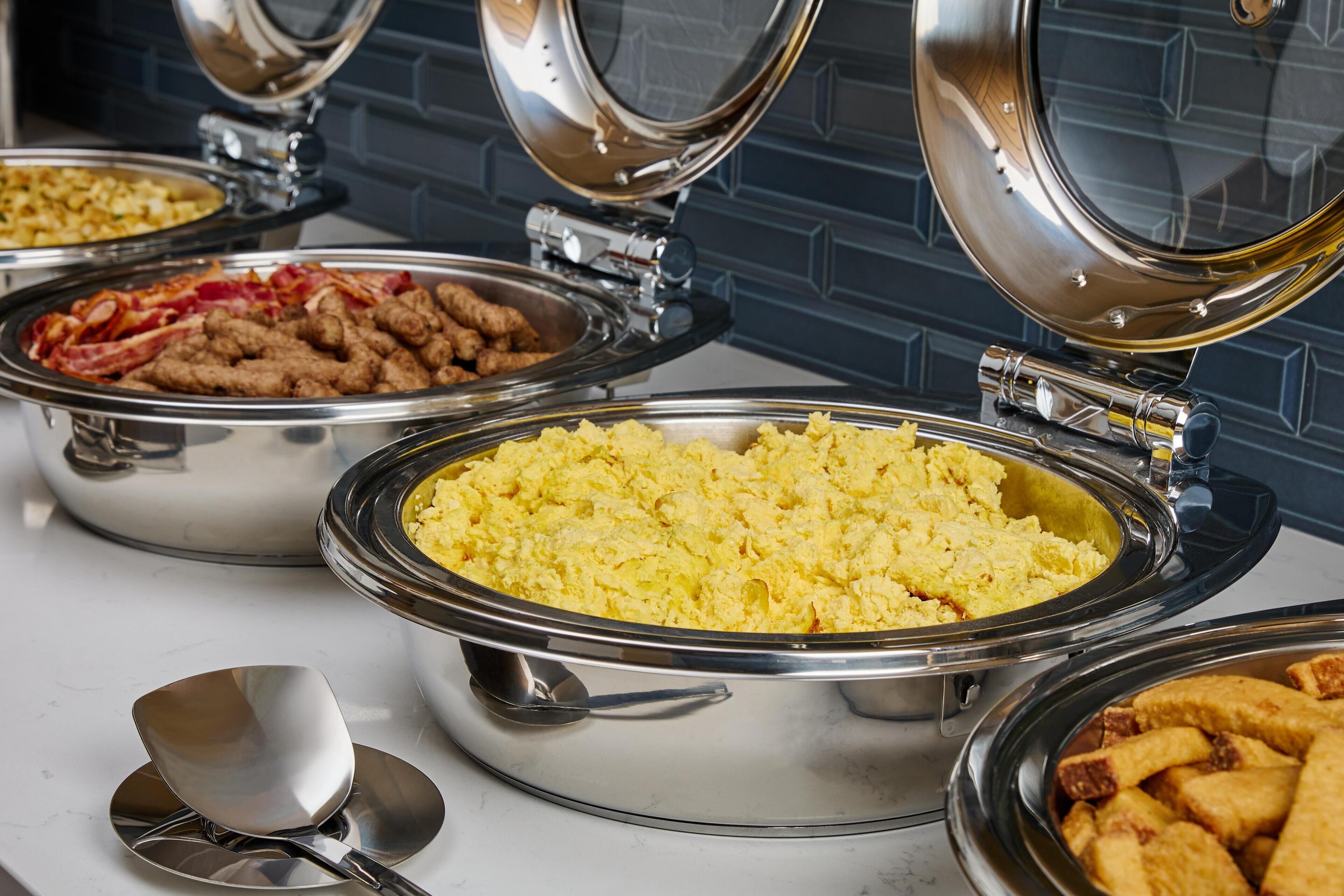 Buffet com diversos alimentos em recipientes de metal, incluindo ovos mexidos, bacon e batatas fritas.