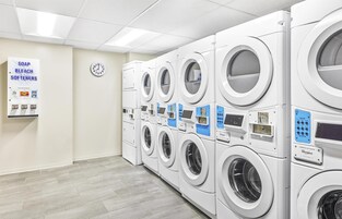 A laundromat with multiple washers and dryers, a clock, and signs for soap, bleach, and softeners.