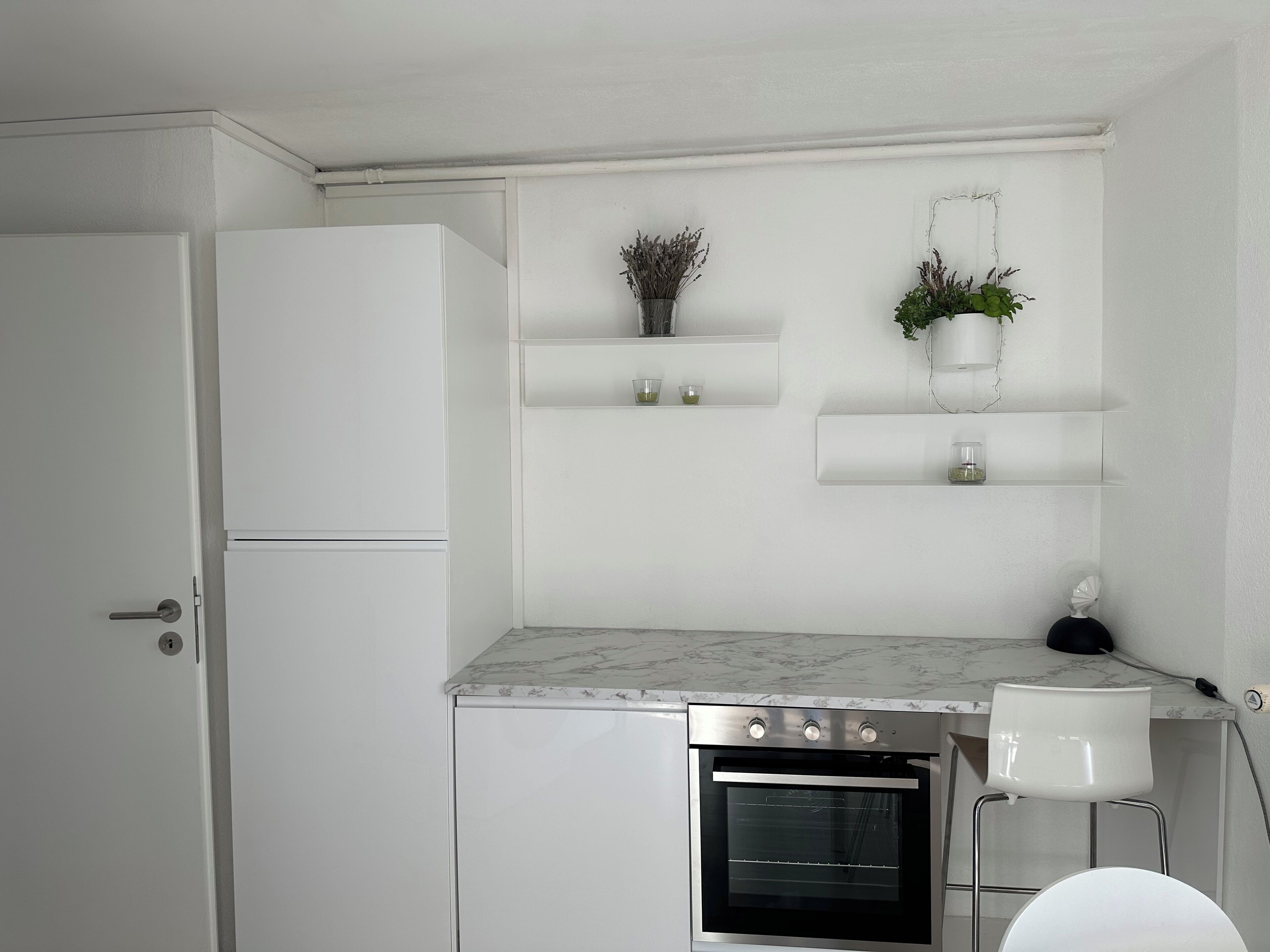 Cozinha compacta com armários brancos, forno embutido e bancada de mármore. Há duas prateleiras com plantas e recipientes de vidro.