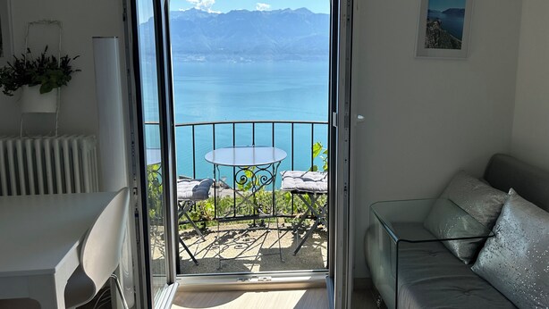 Ein Balkon mit Tisch und Stühlen, einer Couch und Blick auf einen See und Berge.