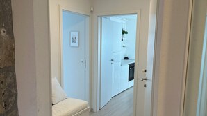 Ein Flur, der zu einem Schlafzimmer führt, mit einem Bett, einem Heizkörper und einem durch eine offene Tür einsehbarer Küchenbereich.